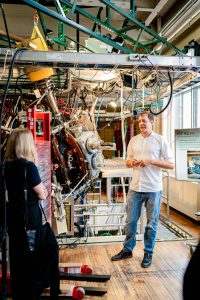 A man in a white dress shirt and jeans stands next to a large piece of scientific equipment and speaks to an audience.