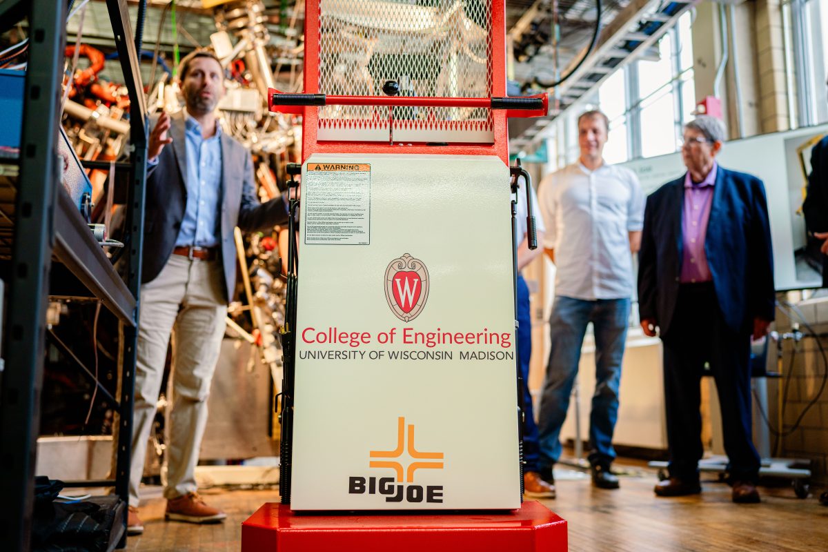 A small forklift sits in a well-lit room, surrounded by people and scientific equipment. The front reads "College of Engineering: University of Wisconsin-Madison."