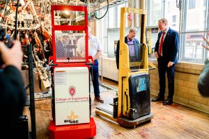 Two small forklifts sit in a well-lit room, surrounded by people and scientific equipment.