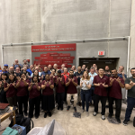 A group of people stand in a concrete testing lab and hold their hands up to form Ws.