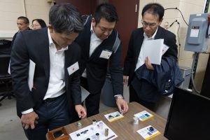 Three Asian men bend over a table to look at small 3D printed parts