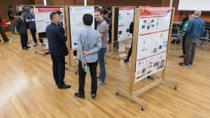 Three men stand in a large room and talk, surrounded by large research posters
