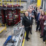 A group of people stands in a large lab space with stairs leading to the main piece of equipment.